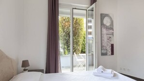 Ein Hotelzimmer mit Bett, Nachttisch, Lampe, Fenster mit Vorhängen und Blick auf einen Balkon mit Stuhl und Tisch.