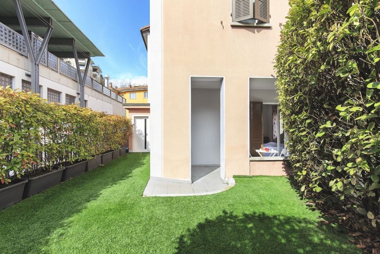 Un edificio moderno con techo plano, un pequeño patio con césped artificial y un seto alto.
