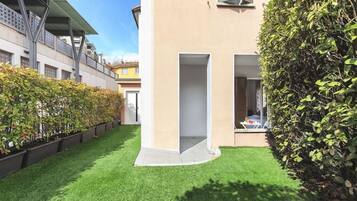 Un edificio moderno con techo plano, un pequeño patio con césped artificial y un seto alto.