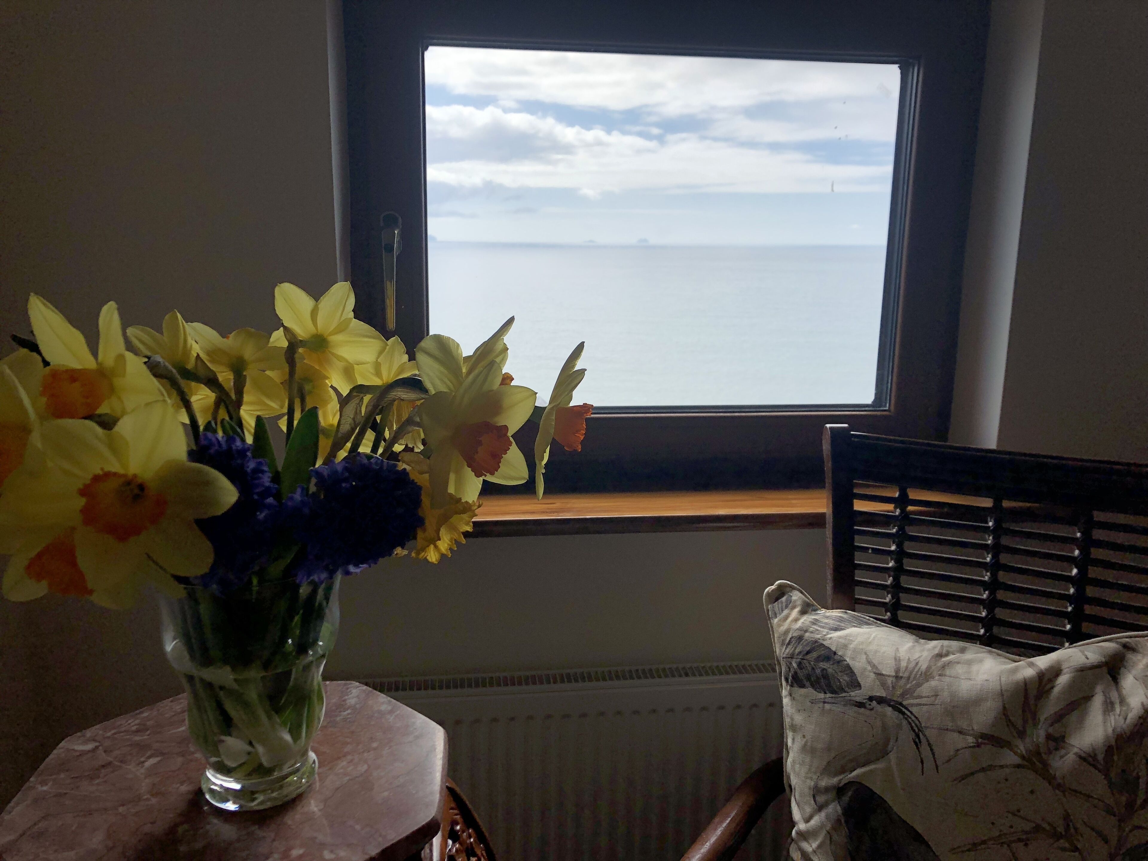 A window with a view of the sea, a vase with yellow and purple flowers on a table, and a chair with a patterned pillow.
