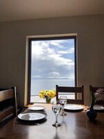 Una mesa de comedor con platos, vasos y un florero, frente a una ventana con vistas al mar y nubes.
