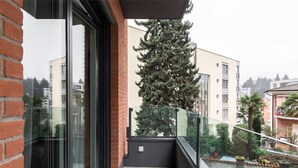 A balcony with a glass railing, a brick wall, and a view of a tall evergreen tree. - Bright one-bedroom apartment 5 minutes from the center - MONTARINA (Lugano)