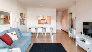 Un salon moderne avec un canapé, un coin repas avec des chaises, un téléviseur et une cuisine visible à travers une porte ouverte.