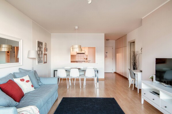 Una sala moderna con sofá, comedor con sillas, televisor y cocina visible a través de una puerta abierta.