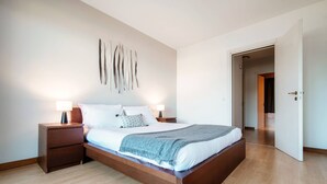 Une chambre moderne avec un lit en bois, du linge de lit blanc et un plaid gris. On y trouve deux tables de chevet avec des lampes, un luminaire fixé au mur et une porte donnant sur une autre pièce.