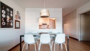Un espace repas moderne avec une table en verre et des chaises blanches, un luminaire suspendu et une cuisine visible à travers une porte ouverte.