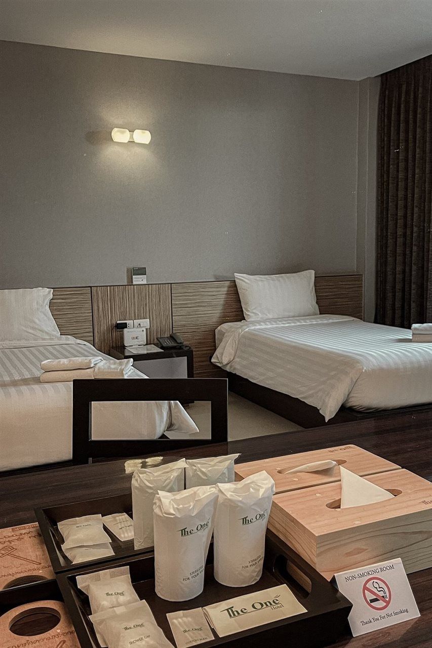 Habitación de hotel con dos camas, una mesita de noche con teléfono y reloj, y un atril con artículos de aseo y un cartel de no fumar.