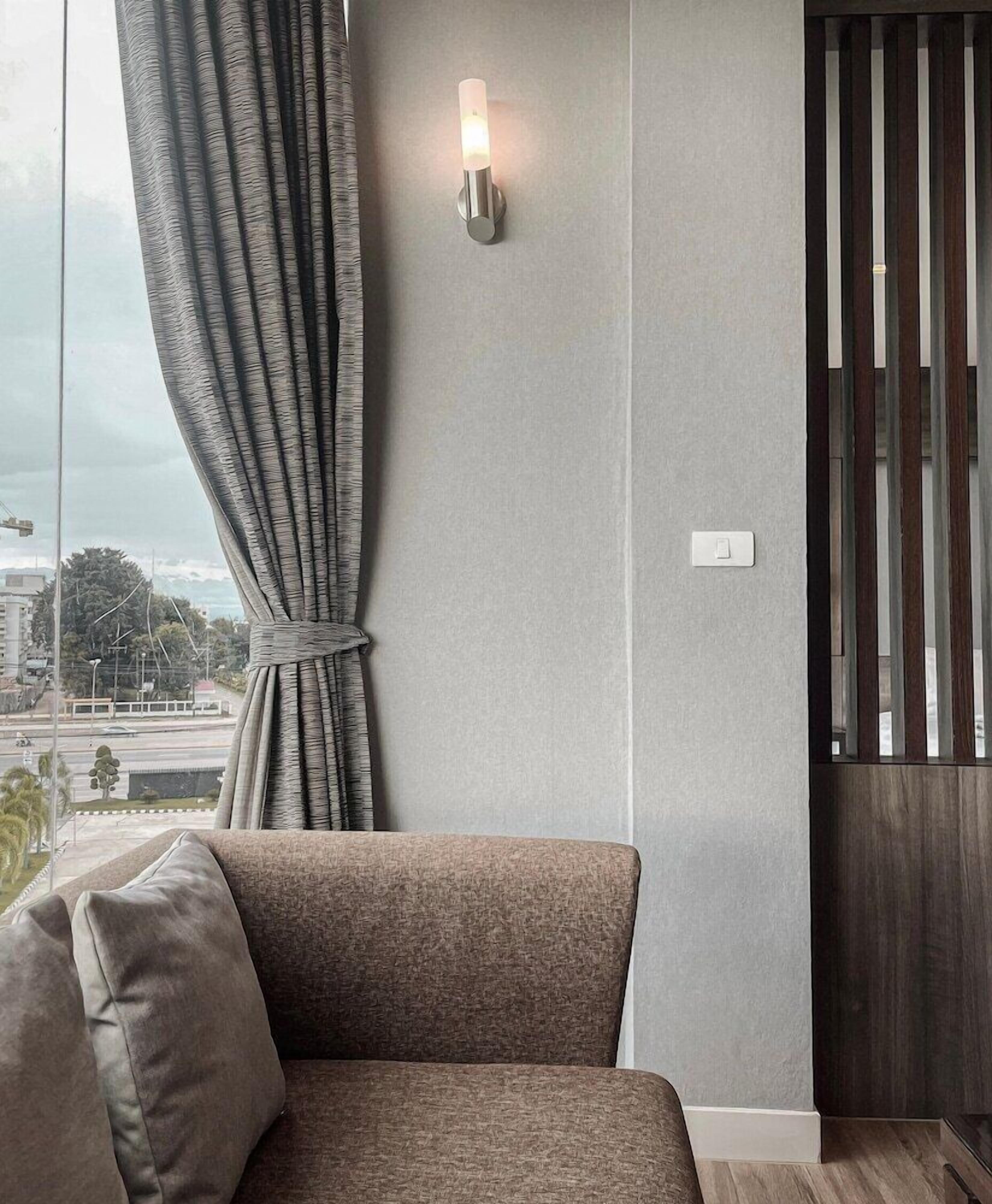 Un salon moderne avec un canapé gris, un luminaire fixé au mur et une vue sur le paysage urbain, visible à travers une fenêtre ornée de rideaux gris.