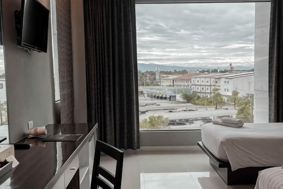Ett sovrum med ett stort fönster, en säng, en tv som är monterad på väggen och ett skrivbord med en stol.