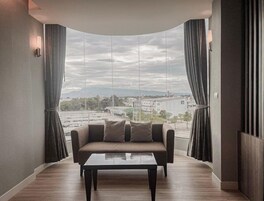 Uma sala de estar moderna com sofá e mesa de centro, janelas amplas com cortinas e vista para o horizonte da cidade.