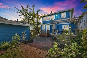 A residential backyard with a brick pathway, a wooden deck, and a blue house.