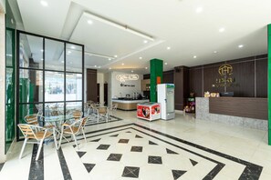 A hotel lobby with a reception desk, a coffee and tea station, and a dining area with chairs and tables.
