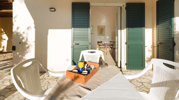 Un espace extérieur abrité, aménagé avec une table et des chaises, une boîte en bois contenant des bocaux, et une vue sur une pièce dotée d’une porte et de fenêtres à volets.