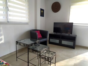 A living room with a glass-top table, a black sofa with a red pillow, a TV on a black entertainment unit, and a wall-mounted decorative piece.