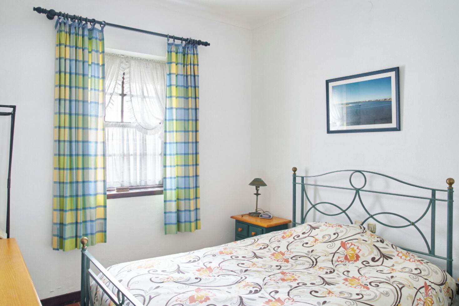 A bedroom with a bed, a window with curtains, a bedside table with a lamp, and a framed picture on the wall.