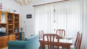 Uma sala de estar com sofá azul, mesa de jantar de madeira e cadeiras, televisão e estante com diversos objetos.
