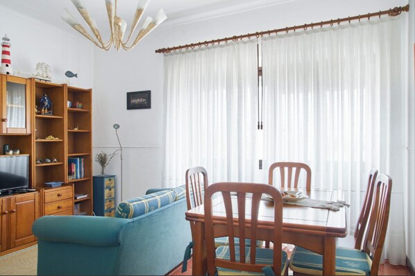 Una sala de estar con un sofá azul, una mesa de comedor de madera y sillas, un televisor y una estantería con diversos objetos.