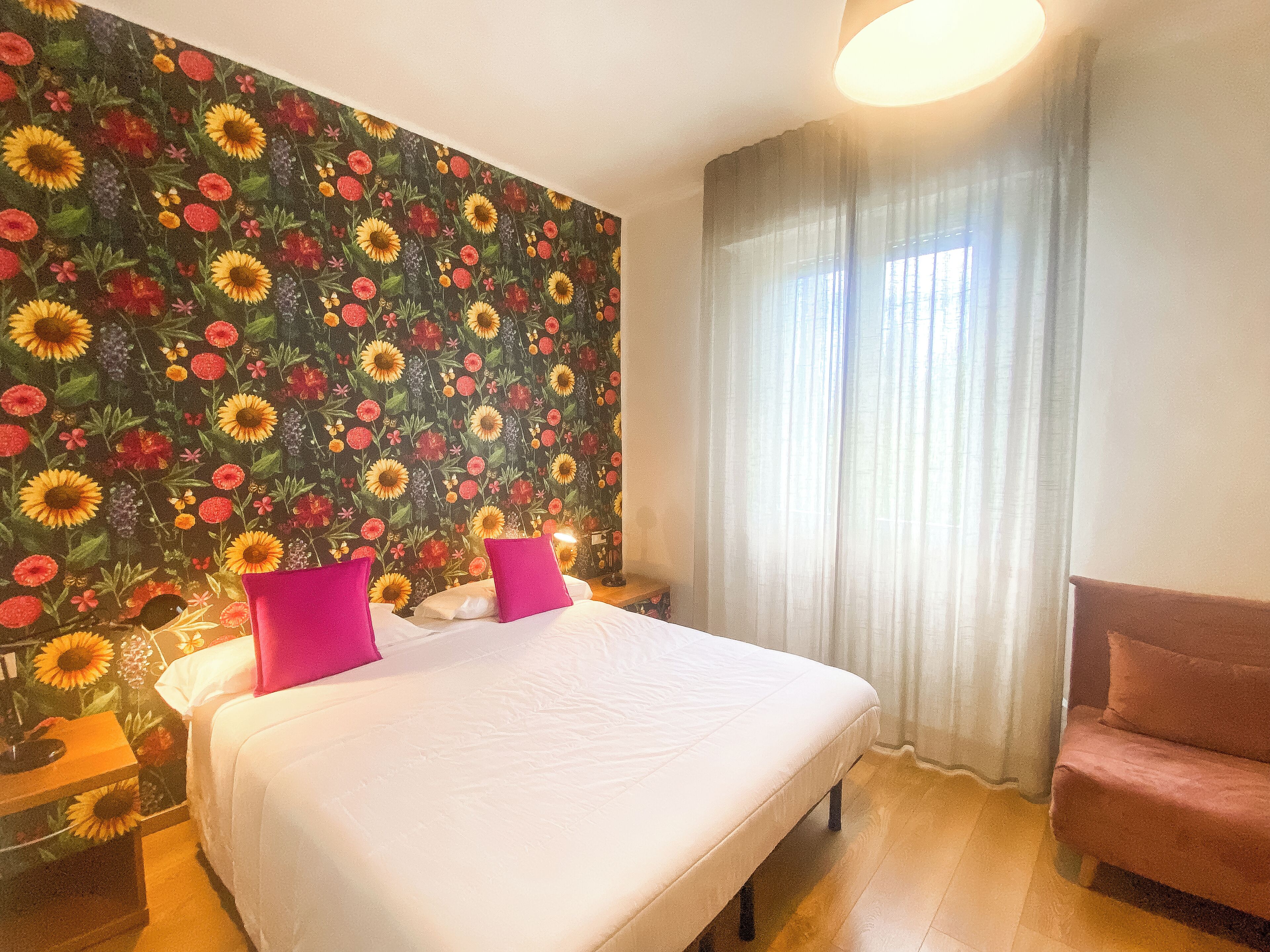 Una habitación de hotel con una cama grande, dos almohadas rosas, una mesita de noche de madera, una lámpara, una ventana con cortinas de encaje y un papel tapiz floral.