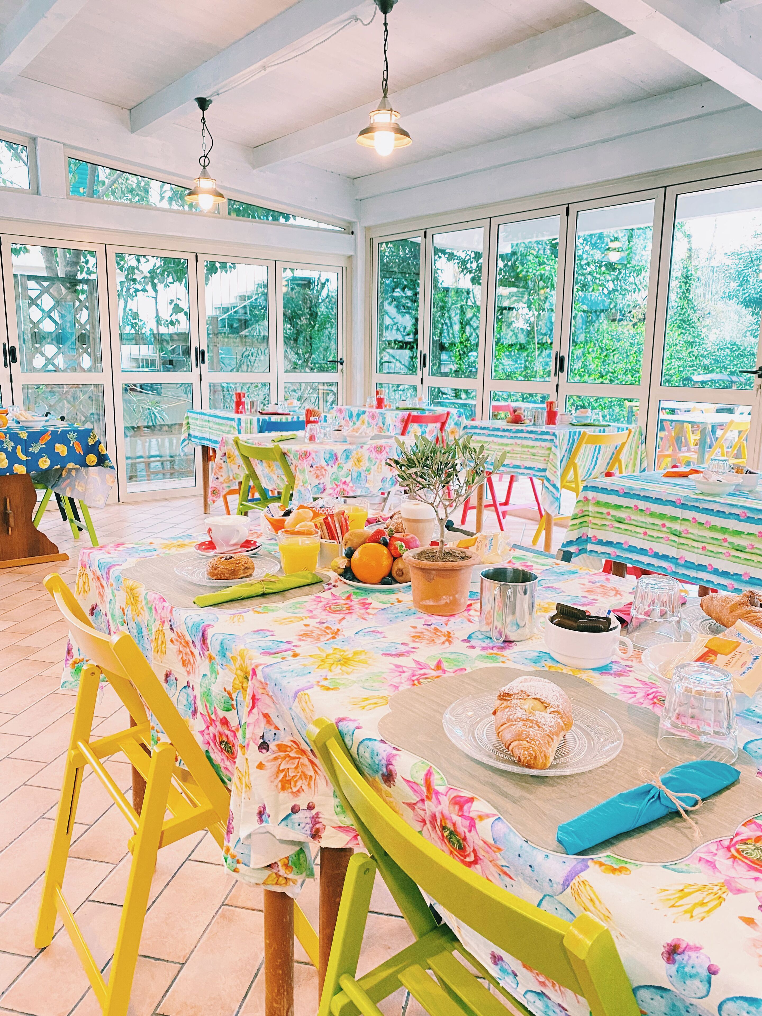 Un comedor luminoso con ventanas amplias, una mesa dispuesta con comida y bebidas, y sillas coloridas.