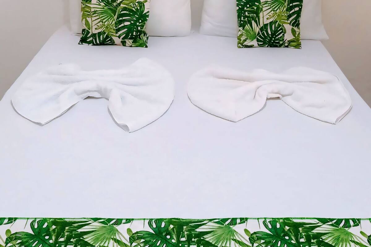 Un lit avec du linge de lit blanc, deux oreillers ornés d’un motif de feuilles tropicales et, au-dessus, une œuvre d’art aux couleurs rouge et jaune.