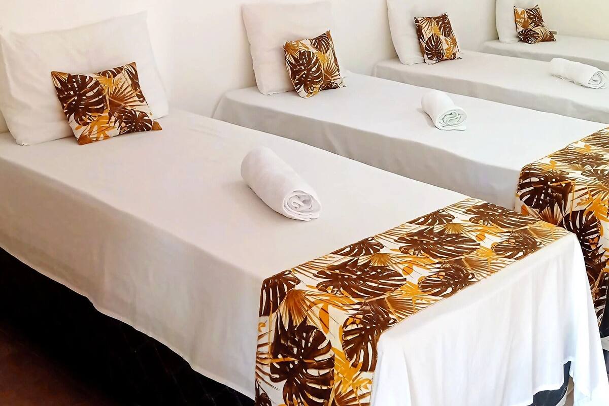 Une chambre d’hôtel avec deux lits simples, chacun recouvert d’une courtepointe blanche et d’un plaid à motifs tropicaux. Un mur est peint dans une couleur vive, et une hélice de ventilateur est fixée au plafond.