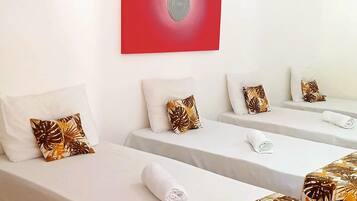 A hotel room with two single beds, each with a white bedspread and a tropical leaf-patterned throw. There is a red accent wall and a ceiling fan.