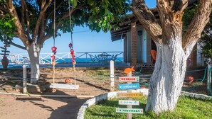 Une zone située au bord de la plage, avec un panneau indicateur en bois portant plusieurs flèches directionnelles, un arbre, et une vue sur la mer en arrière-plan.