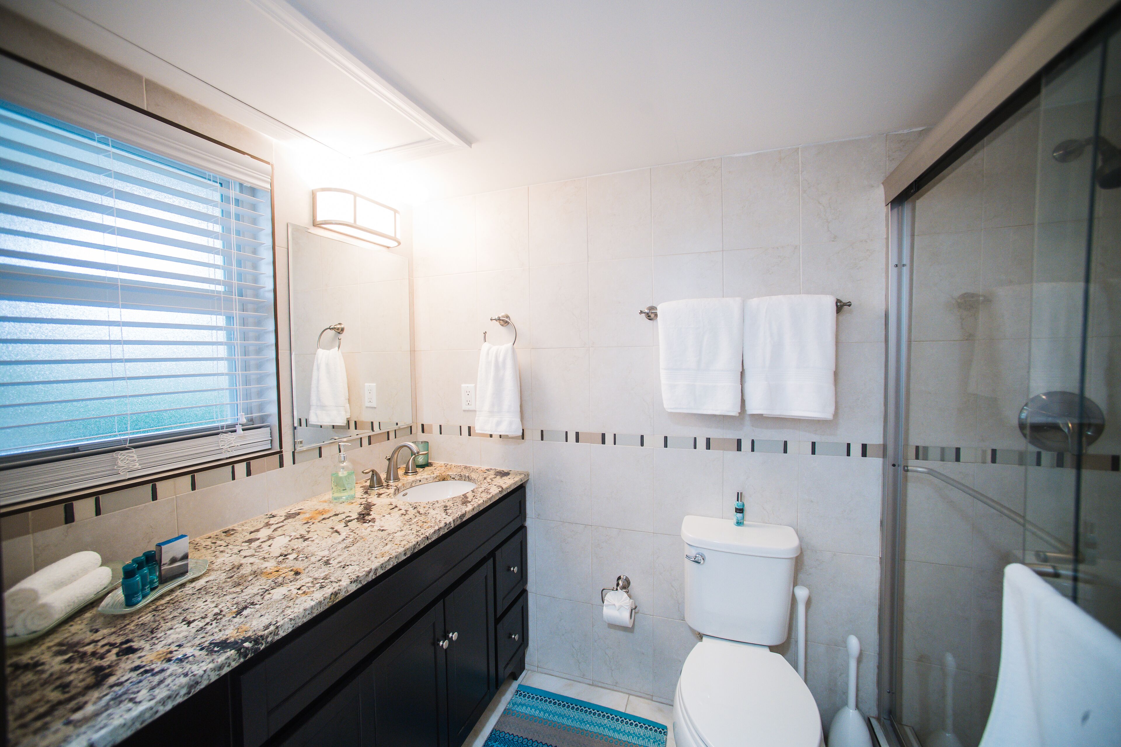 A modern bathroom with a granite countertop, dark cabinets, a toilet, a glass shower enclosure, and a window with blinds.