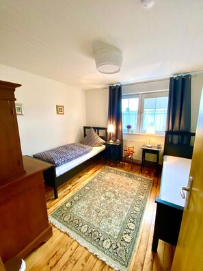 Een kleine, knusse kamer met een bed, een bureau, een stoel, een radiator en een raam met gordijnen.