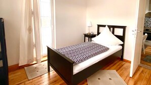 Une chambre avec un plancher en bois, un lit recouvert d’une couvre-lit à motifs, une table de chevet avec une lampe et une fenêtre avec des rideaux.