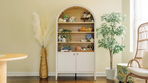 Um quarto com uma estante branca em arco, uma cadeira de vime, uma planta em vaso e uma mesa de madeira redonda.