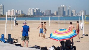 Uma quadra de voleibol de praia com jogadores, um guarda-sol colorido e um horizonte urbano ao fundo.