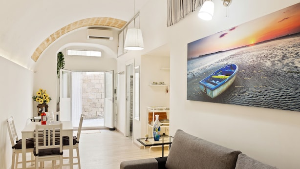 A modern living room with a large painting of a sunset over the sea, a grey sofa, and a dining area with a white table and chairs. - San Nicola Casa Clemente Apartment (Bari)