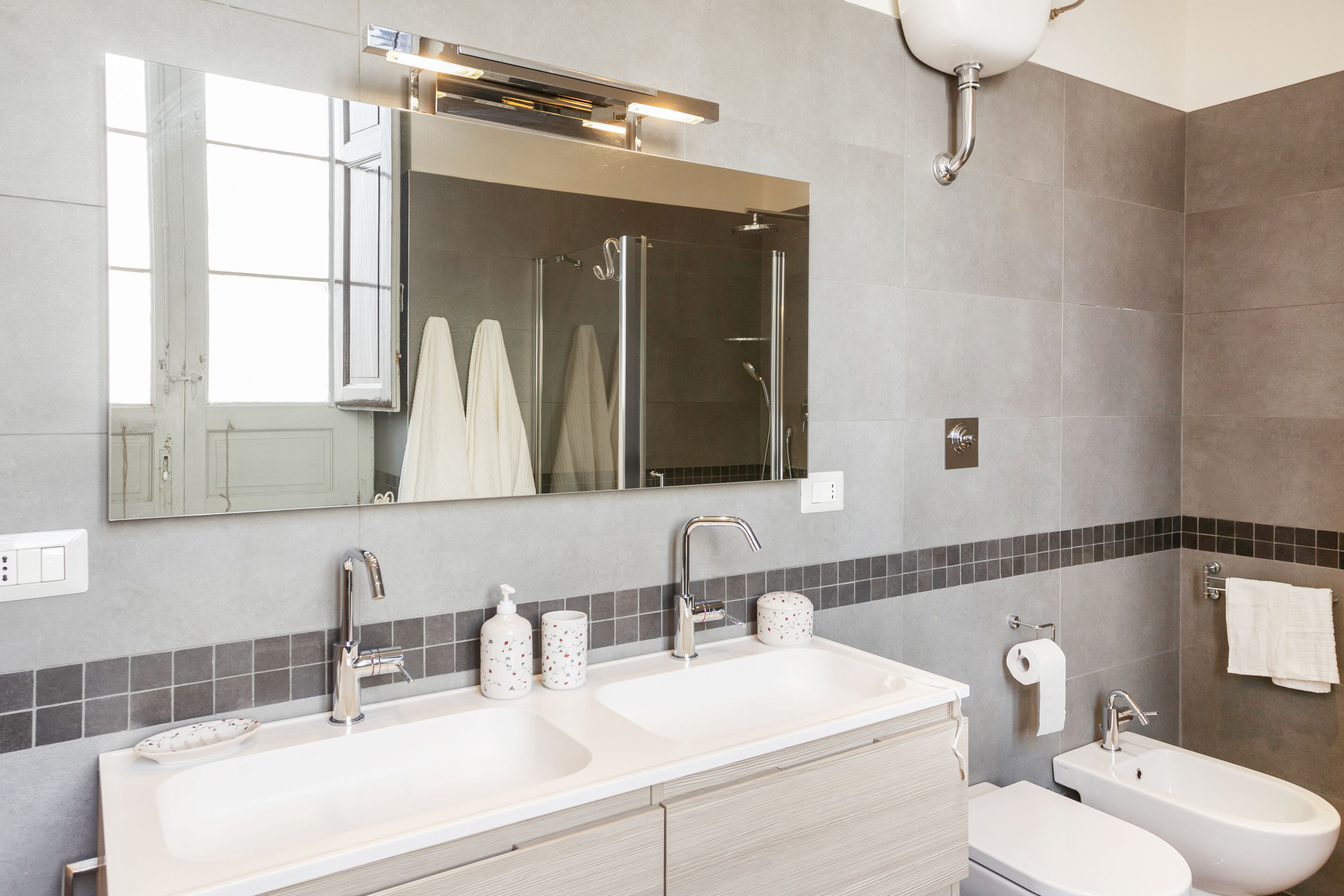 Un baño moderno con dos lavamanos, ducha y bidé. Incluye un espejo grande, un perchero para toallas y un dispensador de papel higiénico.