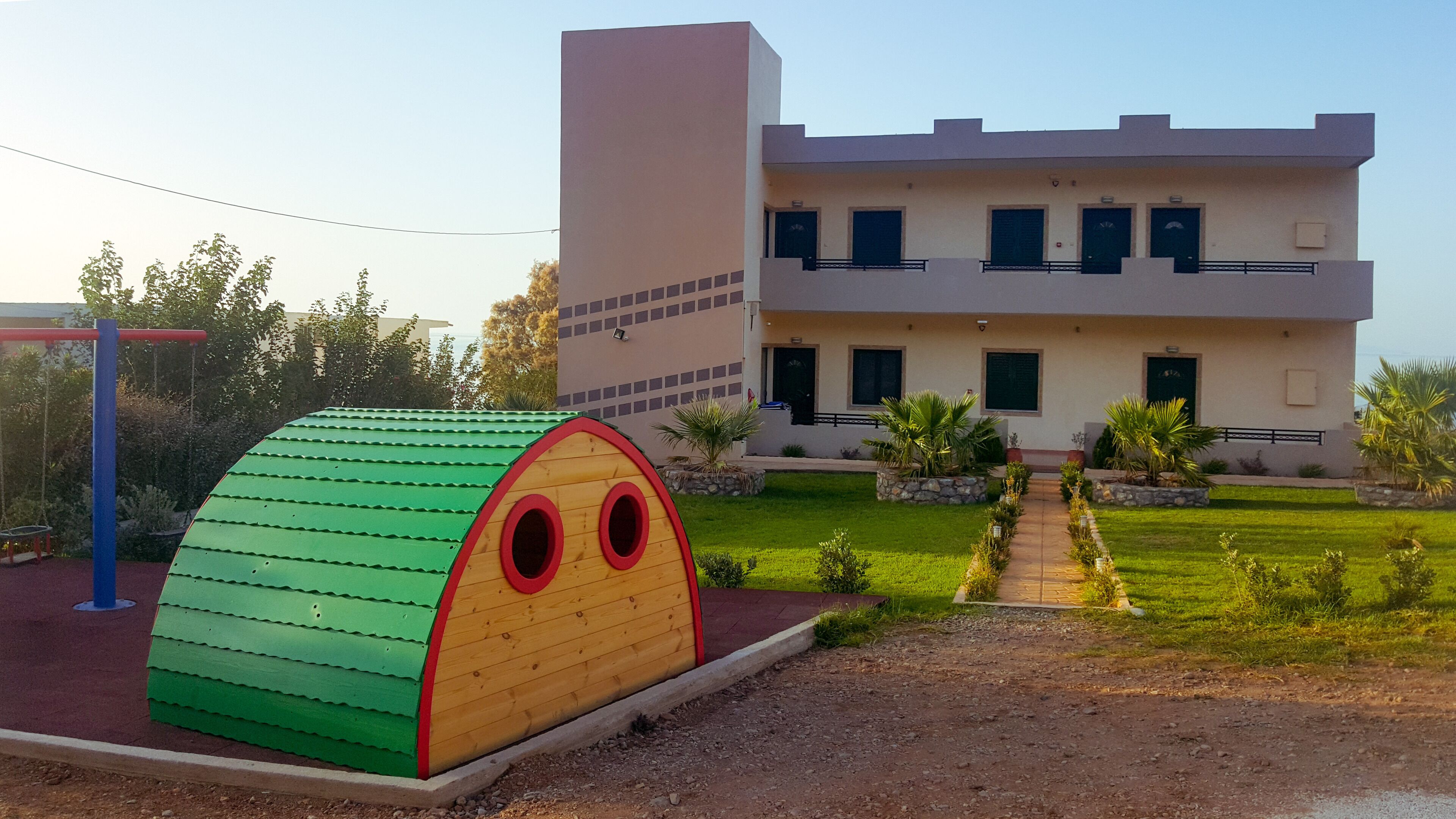 A modern building with a flat roof and a symmetrical facade, a green play structure resembling a house, and a well-maintained lawn with a pathway leading to the building.