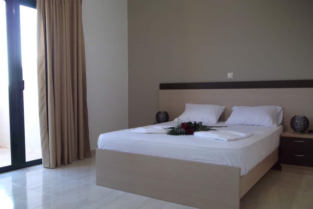 A hotel room with a large bed, a nightstand, a lamp, and a view of the outside.