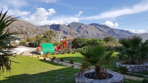 Un parque infantil con tobogán y estructuras para escalar, rodeado de césped y palmeras, con montañas al fondo.