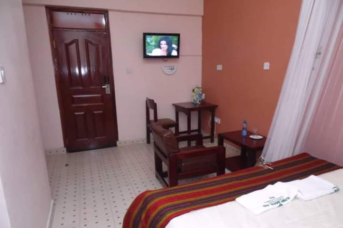 A hotel room with a bed, a TV mounted on the wall, a wooden door, and a small table with a vase.