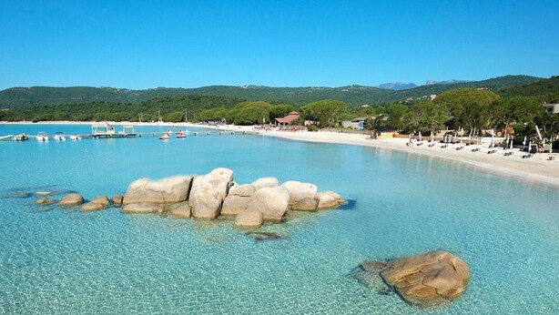 Una playa costera con aguas cristalinas, rocas grandes en el centro y una orilla arenosa con sillas y sombrillas de playa.