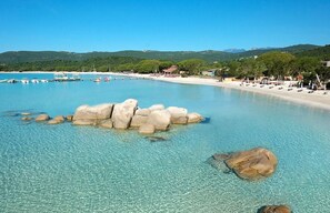 Een kuststrand met helder water, grote rotsen in het midden en een zandstrand met ligbedden en parasols.