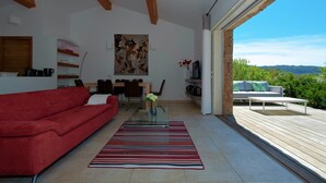 Una sala de estar moderna con un sofá rojo, una mesa de centro de vidrio y un tapete a rayas. Hay un comedor con una mesa y sillas, y un cuadro en la pared. La habitación tiene un amplio ventanal corredero que da a una terraza exterior con vistas a zonas verdes.