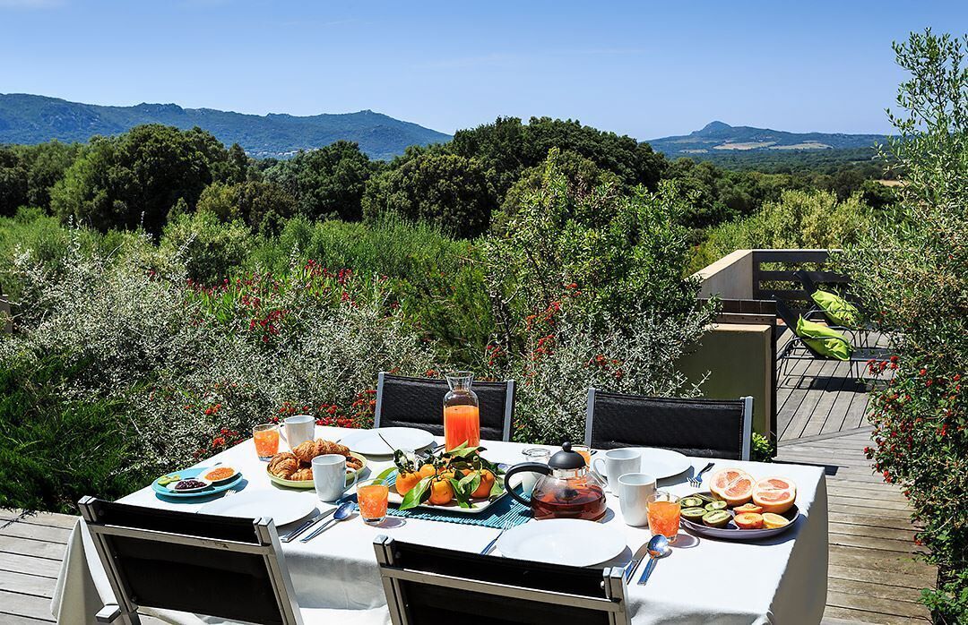 An outdoor dining area with a table set for breakfast, surrounded by lush greenery and mountains in the background.
