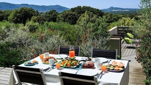Un área de comedor al aire libre con una mesa dispuesta para el desayuno, rodeada de abundante vegetación y montañas al fondo.