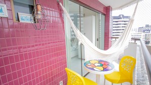 A balcony with pink tiled walls, a hammock, a round table with a colorful mosaic top, and two yellow chairs. - PB02 Ap. a 500m do Fiesta Pituba (Salvador)