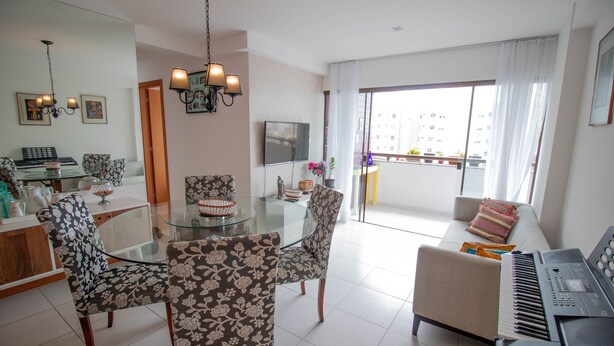 A modern living room with a glass dining table, floral patterned chairs, a sofa, a piano, and a balcony with a view of buildings. - PB02 Ap. a 500m do Fiesta Pituba (Salvador)
