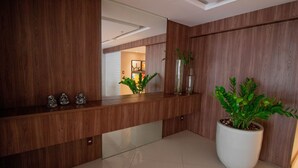 A reception area with a wooden reception desk, a large potted plant, and a mirror reflecting the interior. - PB02 Ap. a 500m do Fiesta Pituba (Salvador)