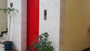 A hallway with a red door, a potted plant, and a wall-mounted elevator control panel. - Avenue Suites Hotel (Nakuru)