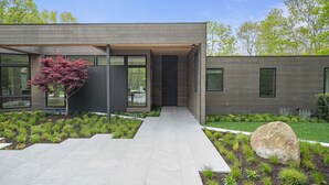 Una casa moderna con un porche cubierto, ventanas amplias y un jardín ajardinado con una roca destacada y un árbol de flores rosadas.