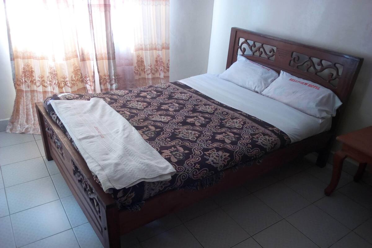 A neatly made bed with a patterned bedspread and a white pillow, a wooden headboard with intricate carvings, and a small wooden table beside the bed.