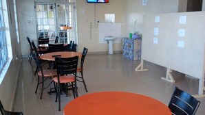 A dining area with round orange tables and black chairs, a television mounted on the wall, and a window with a view of the outside. - Bellview Hotel (Thika)
